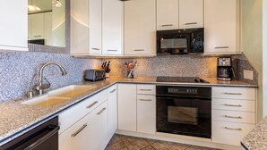 A modern kitchen with white cabinets, a granite countertop, a built-in oven, a microwave, and a coffee maker. - Beachfront Living - Makena Surf E-202 - Mesmerizing Views & Tropical Landscapes (Makena, Maui)