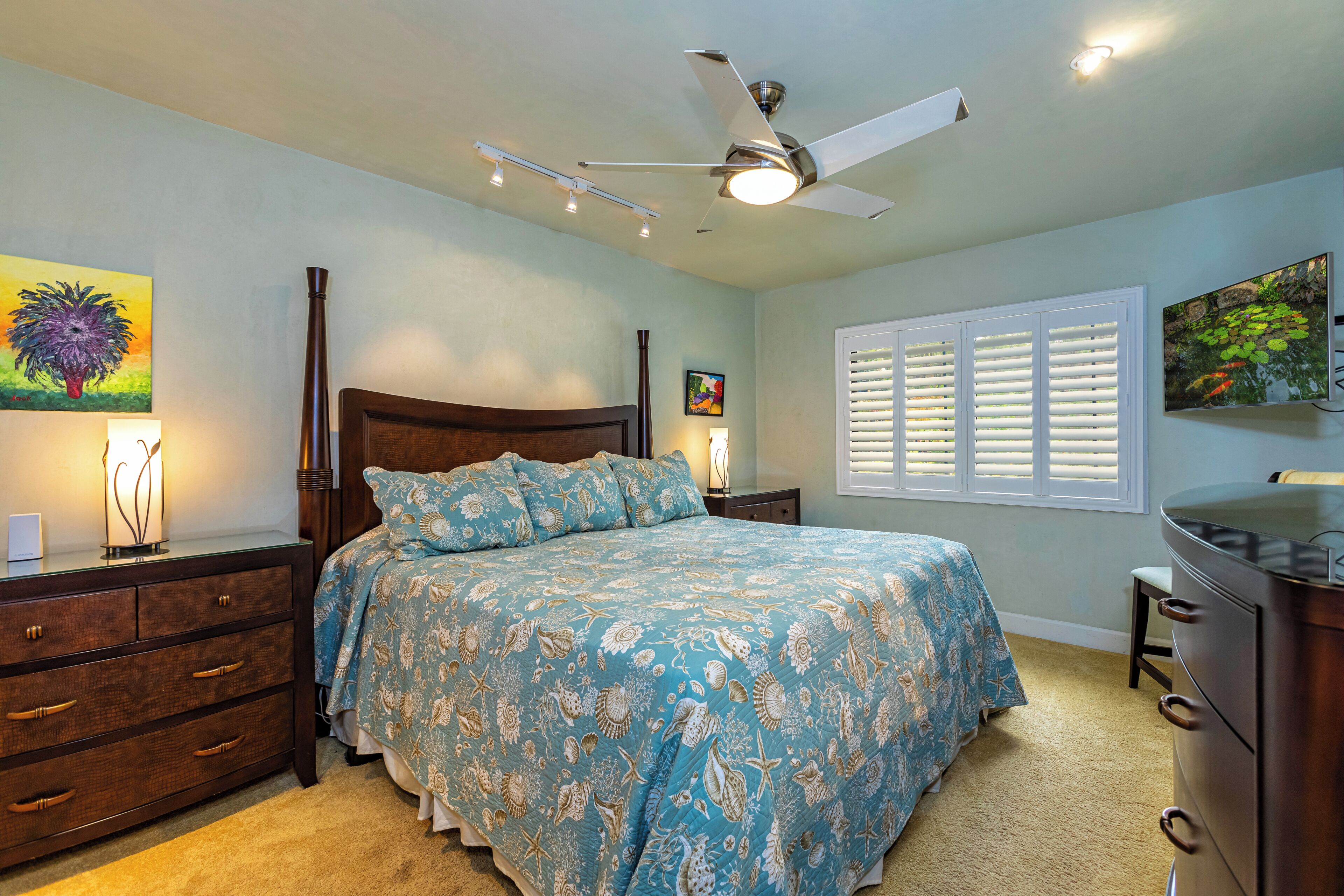 Une chambre à coucher comprenant un lit, une commode, une table de chevet, un ventilateur de plafond et des œuvres d’art accrochées aux murs.
