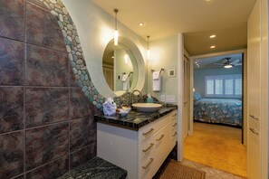 Un baño con pared de piedra, un lavamanos y un espejo. Se ve una cama al fondo.