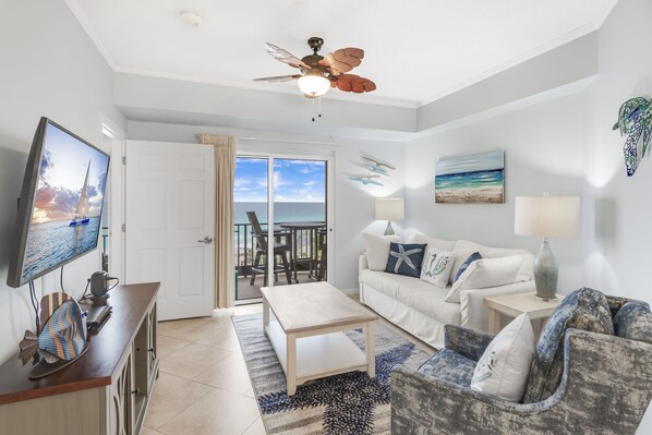 A modern living room with a television, a ceiling fan, a sofa, a coffee table, and a view of the ocean through a sliding glass door. - 6-Seat Golf Cart ~ Beach Front ~ Private Balcony (Miramar Beach)