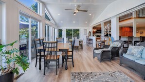 A spacious living area with a dining table, chairs, and a sofa. There is a ceiling fan, large windows, and a view of the outdoors.