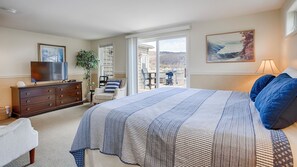 Un dormitorio con una cama grande, un aparador, un televisor, una silla, un balcón con vistas y un cuadro en la pared.