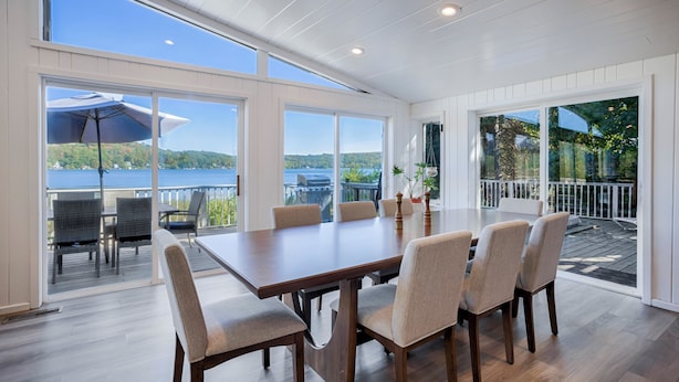 A dining area with a large table and chairs, large windows offering a view of a lake and hills, and a deck with outdoor seating.