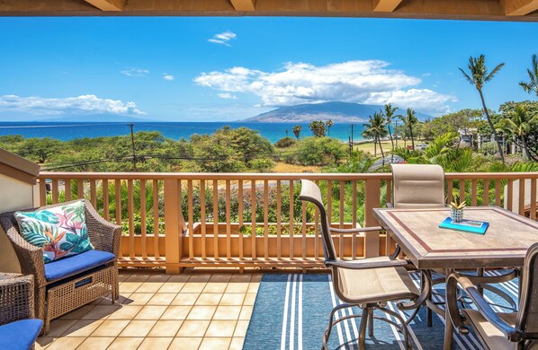 A balcony with wicker furniture, a table, and a view of the ocean and a mountain.
