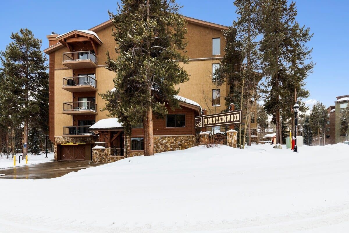 A multi-story building with a sign reading "Mountain Wood." It features balconies, a stone facade, and is surrounded by trees and snow.
