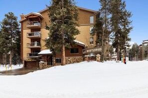 Un immeuble à plusieurs étages, sur lequel on peut lire l’inscription « Mountain Wood ». Il est doté de balcons, d’une façade en pierre et est entouré d’arbres et de neige.