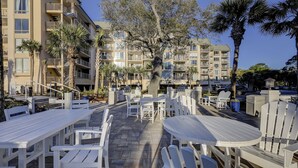 An outdoor patio area with white plastic furniture, including tables and chairs, surrounded by palm trees and a building with multiple balconies.