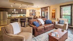 A modern living room with a brown leather sofa, beige armchairs, and a wooden coffee table. A kitchen with dark cabinets and stainless steel appliances is visible in the background.