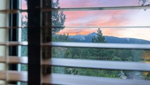 A view through window blinds of a mountainous landscape with trees and a house in the distance.