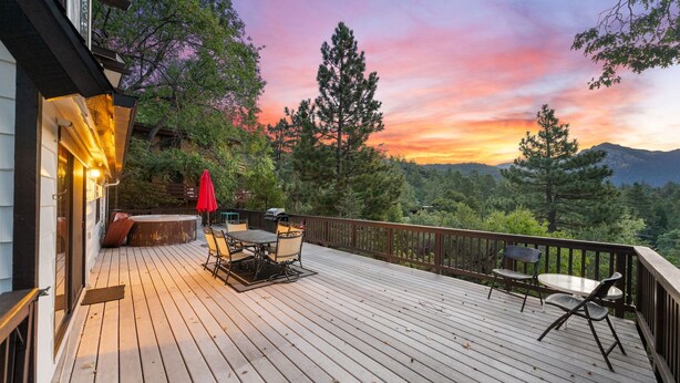 A wooden deck with outdoor furniture, a red umbrella, and a scenic view of trees and mountains during sunset.