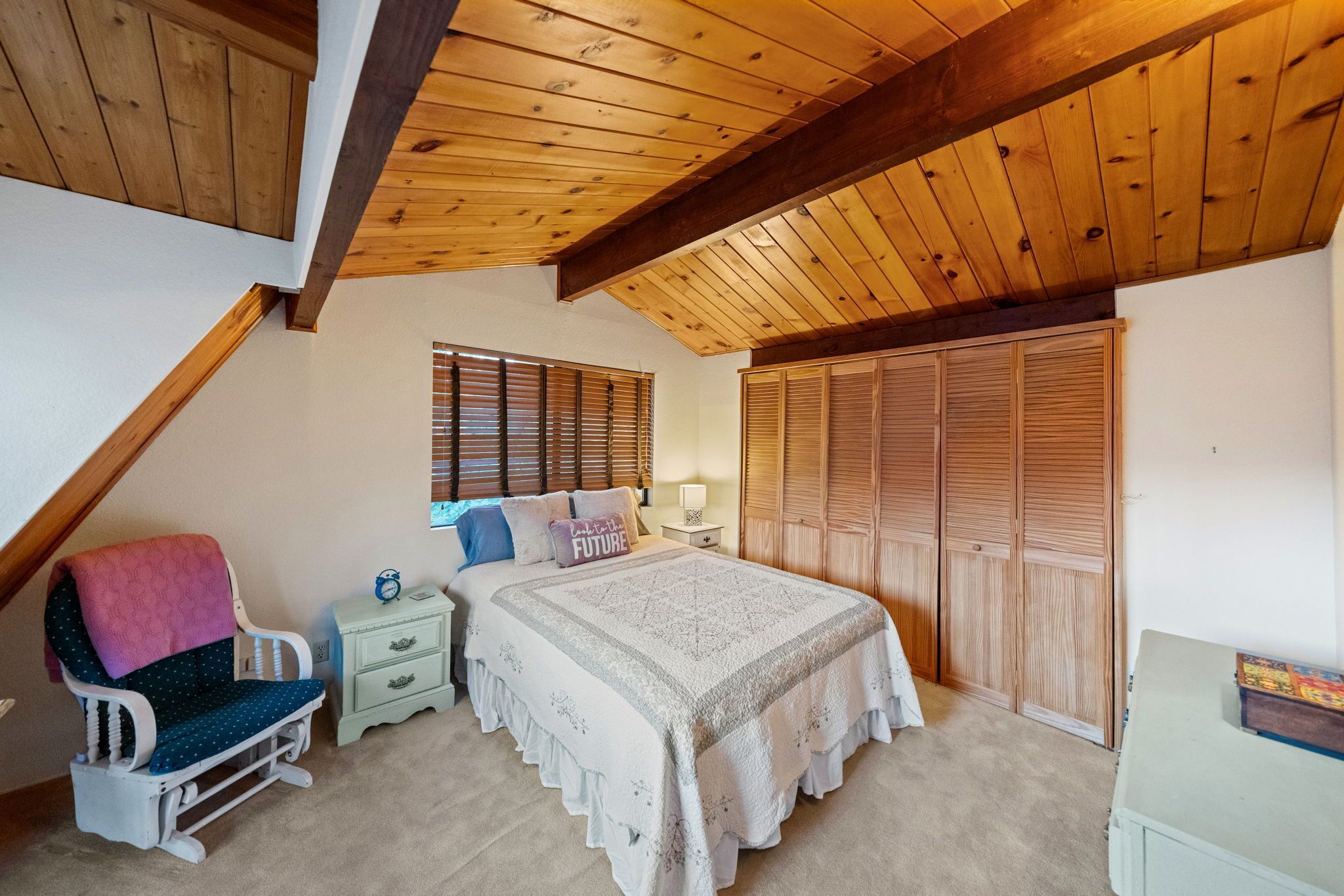 Een slaapkamer met een schuine kap, een bed met een witte spreideken, een schommelstoel, een nachtkastje en een houten garderobe.