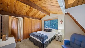 A bedroom with a wooden ceiling, a bed with a blue and white bedspread, a nightstand with a lamp, a window with a cross on the wall, and a blue chair.