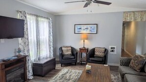 Una sala de estar con ventilador de techo, un televisor de pantalla plana montado en la pared, dos sillones con cojines estampados, una mesa de centro de madera y un sofá con un cojín floral.
