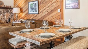 A dining area with a wooden table set for six, a stone wall, a framed picture of a forest, and a poster on the wall. - Beautiful Spacious Pet-Friendly Townhome 264 (Mammoth Lakes)