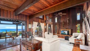 Ein großzügiges Wohnzimmer mit einem weißen Sofa, einem Holztisch und einem Fernseher. Es gibt einen Essbereich mit Stühlen und einem Tisch, sowie einen Blick auf den Ozean durch große Fenster.