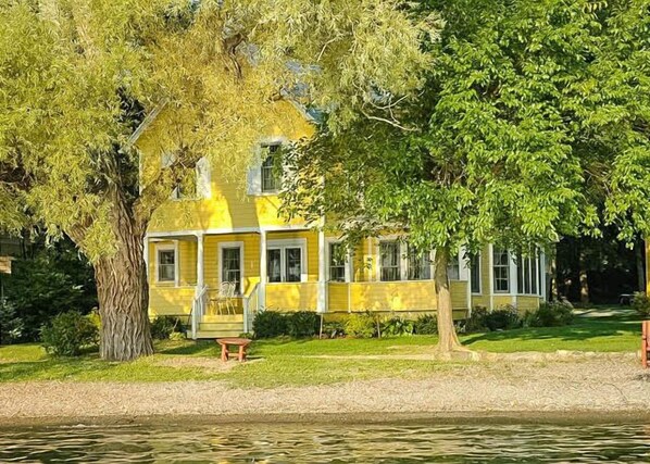 A yellow house with a porch and a bench, surrounded by trees and a body of water. - Heart's Desire: Great Sunset Views (Canandaigua)
