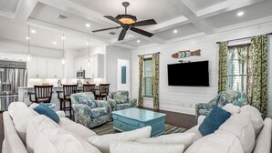 Uma sala de estar moderna com um sofá branco, poltronas estampadas, uma mesa de centro azul e um ventilador de teto.