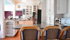 A modern kitchen with white cabinets, a central island, and a built-in oven. There are wicker chairs with a geometric pattern and a toaster oven on the counter. - Aurora Escape: "Spectacular Design Awaits You!" (Aurora)