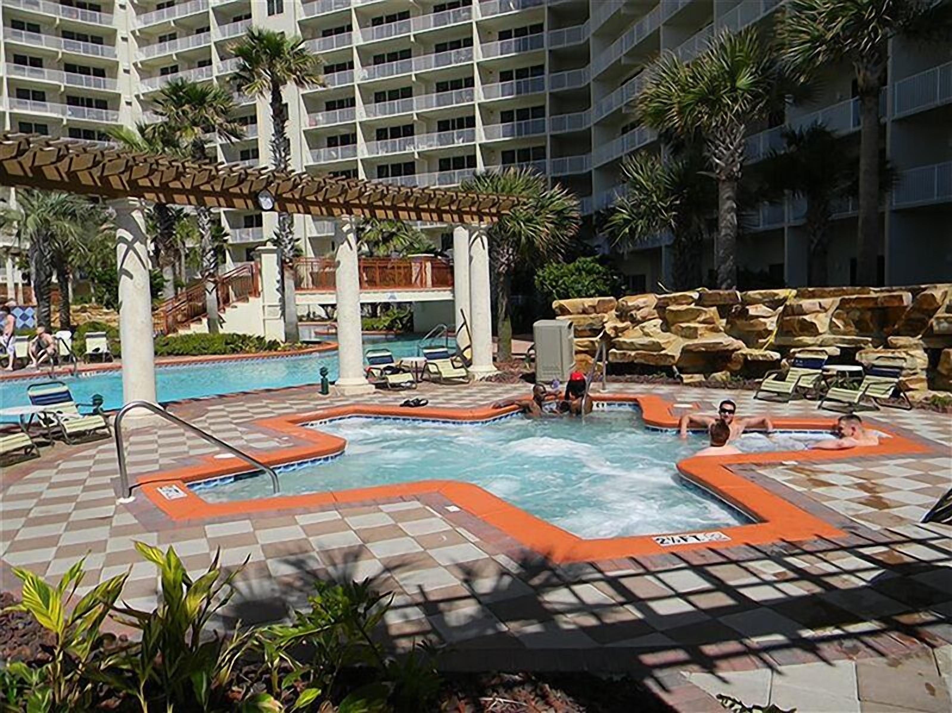 Un área de piscina con varias fuentes de agua, que incluye un jacuzzi y una piscina con olas artificiales, rodeada de sillones de descanso y palmeras.