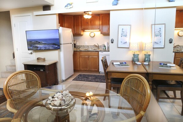 A compact kitchen with wooden cabinets, a refrigerator, a dining table with chairs, and a television on the wall.