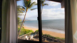 Blick auf einen Strand durch ein Fenster mit Vorhängen, Palmen und einem Geländer.