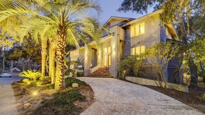Une maison moderne à deux étages, dotée d’une entrée bien éclairée, d’une allée et d’un jardin aménagé avec des palmiers.