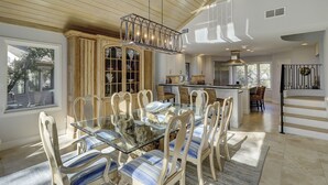 Un vaste espace de salle à manger avec une table en verre et des chaises à rayures, une cuisine équipée d’appareils électroménagers modernes et un escalier doté d’une rampe noire.