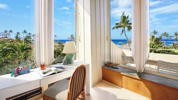 Ein Zimmer mit Schreibtisch, Stuhl und großen Fenstern mit Blick auf einen Strand, an dem Palmen und das Meer zu sehen sind.