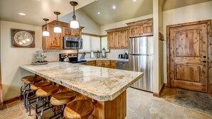 Una cocina con armarios de madera, electrodomésticos de acero inoxidable, una isla de mármol con taburetes altos e iluminación colgante.