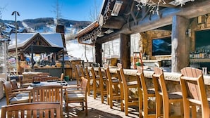 An outdoor seating area with wooden tables and chairs, a bar with a stone wall, and a view of a snowy mountain landscape. - Ritz Carlton 1BR Ski-in/out - 2 Queen Beds - Sleeps 4 (Beaver Creek)