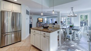 A modern kitchen with a stainless steel refrigerator, white cabinetry, and a granite countertop. There is a dining area with a table and chairs, and a kitchen island with a bowl on the counter. - 106 Oceanwood Trace - 2nd Row Ocean View, Dog Friendly Beachside Oasis. (Hilton Head Island)