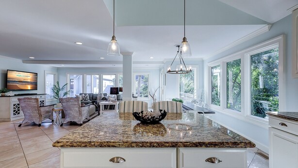 A spacious kitchen with a granite countertop, white cabinetry, and a large island with a decorative bowl. There are pendant lights above the island and a television mounted on the wall. - 106 Oceanwood Trace - 2nd Row Ocean View, Dog Friendly Beachside Oasis. (Hilton Head Island)