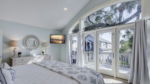 A bedroom with a large bed, a TV, and a view of a house through a glass door. - 106 Oceanwood Trace - 2nd Row Ocean View, Dog Friendly Beachside Oasis. (Hilton Head Island)