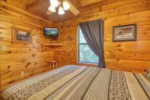 A wooden cabin room with a bed, a television, a window with curtains, and a ceiling fan.