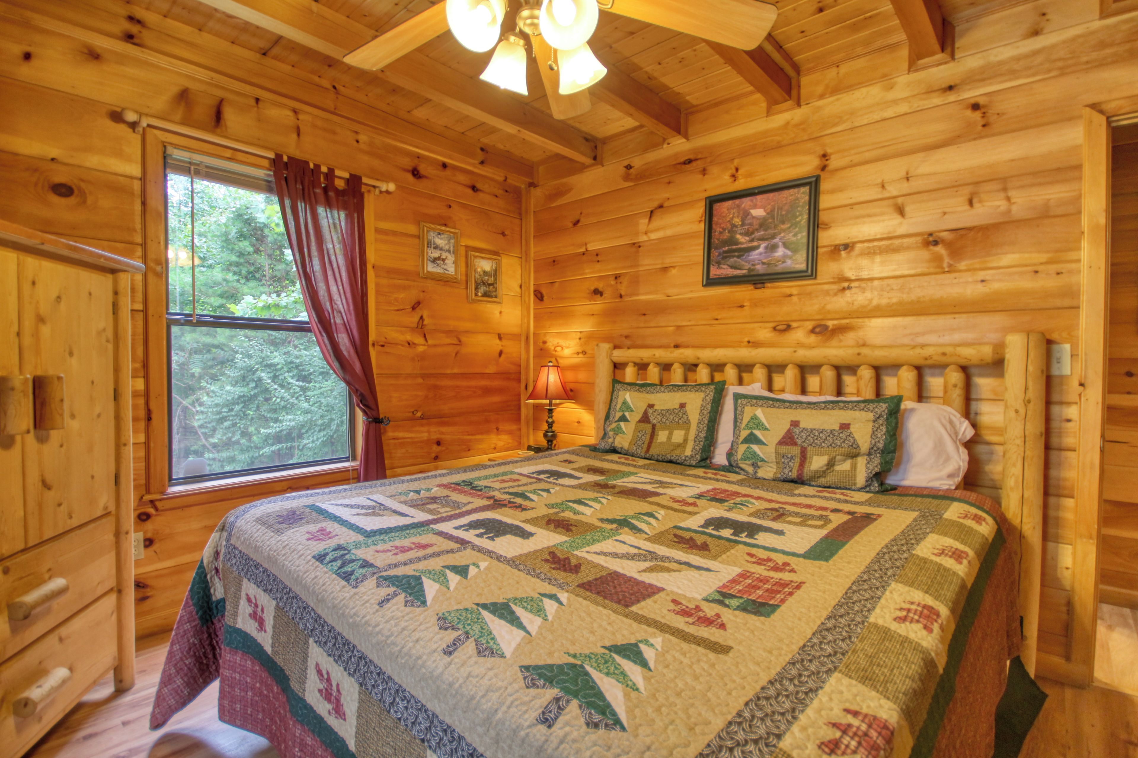 A wooden cabin bedroom with a large bed, a window with curtains, a headboard with pillows, and a ceiling fan.