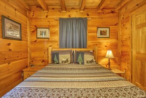 A wooden cabin bedroom with a bed, framed pictures, a lamp, and a window with a curtain. - Privacy~Sunset View~Pet Friendly~Pool Table~Air Hockey (SEVIERVILLE)