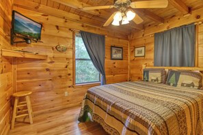 A wooden cabin bedroom with a bed, a ceiling fan, a television, and a window with curtains. - Privacy~Sunset View~Pet Friendly~Pool Table~Air Hockey (SEVIERVILLE)