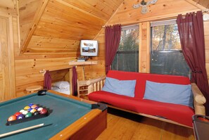 A wooden cabin interior with a pool table, a red sofa with blue cushions, and a television displaying 'Auni Bug's Cabin Rental, LLC'. - Privacy~Sunset View~Pet Friendly~Pool Table~Air Hockey (SEVIERVILLE)