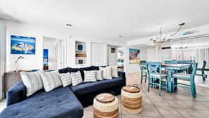 Un salon moderne avec un canapé d’angle bleu foncé, un coin repas avec des chaises bleues et une cuisine en arrière-plan.