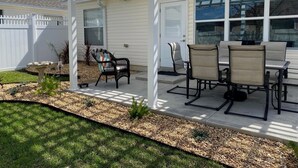 A covered patio with a dining table and chairs, a white fence, and a garden area with a bird bath and plants. - Amazing Location & Beautiful home with Golf Cart, BBQ & WiFi included! (The Villages)
