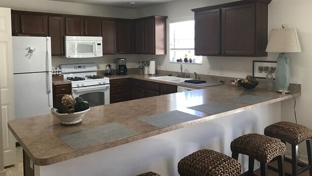 A modern kitchen with dark wood cabinets, a white refrigerator, a microwave, a stove, and a sink. There are bar stools and a lamp on the island. - Amazing Location & Beautiful home with Golf Cart, BBQ & WiFi included! (The Villages)