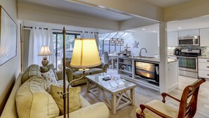 Una sala de estar moderna con una mesa de centro de vidrio, sofá, sillón, chimenea y una cocina con electrodomésticos de acero inoxidable.