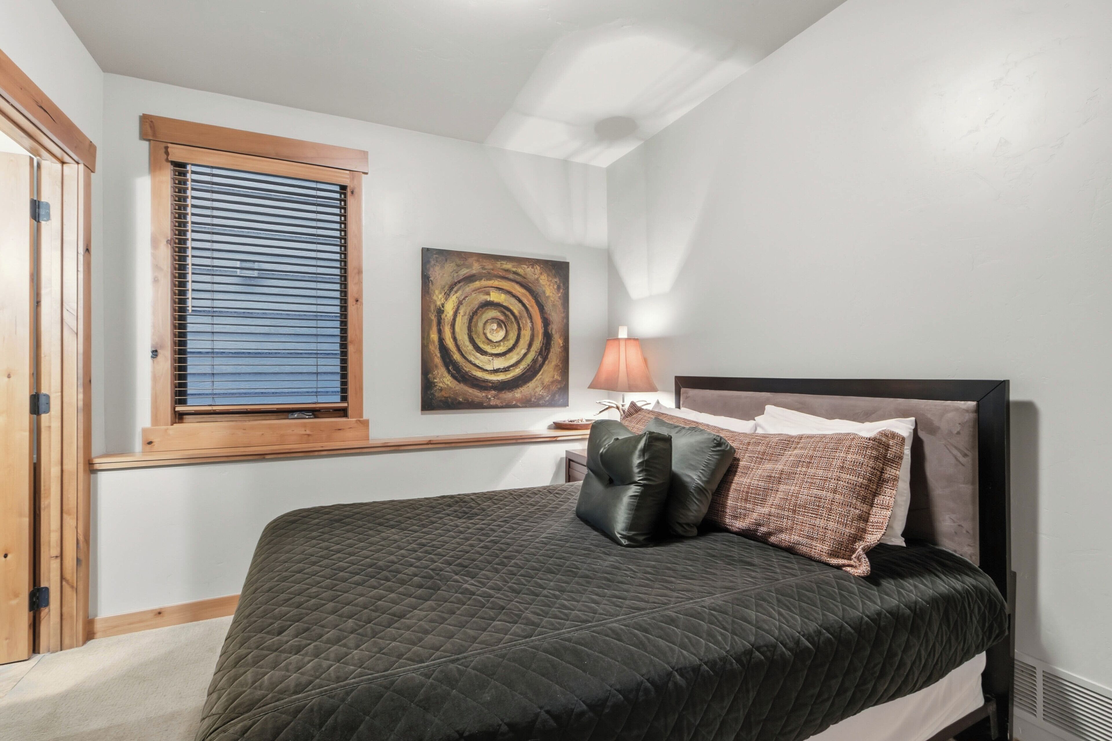 Une chambre à coucher avec un lit, une fenêtre avec des stores, une œuvre d’art accrochée au mur et une lampe de chevet.