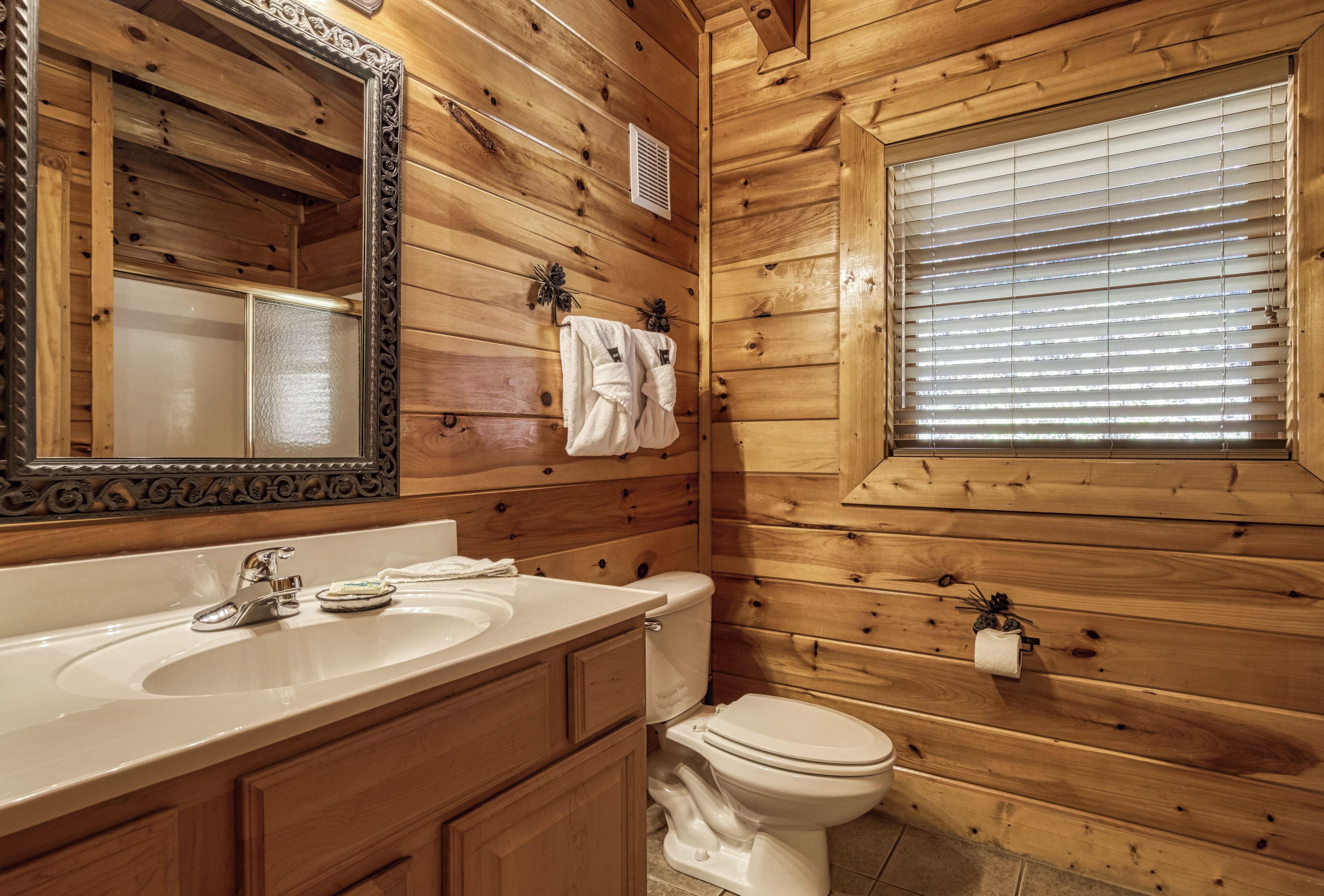 A wooden-paneled bathroom with a large mirror, a sink, a toilet, and a window with blinds.