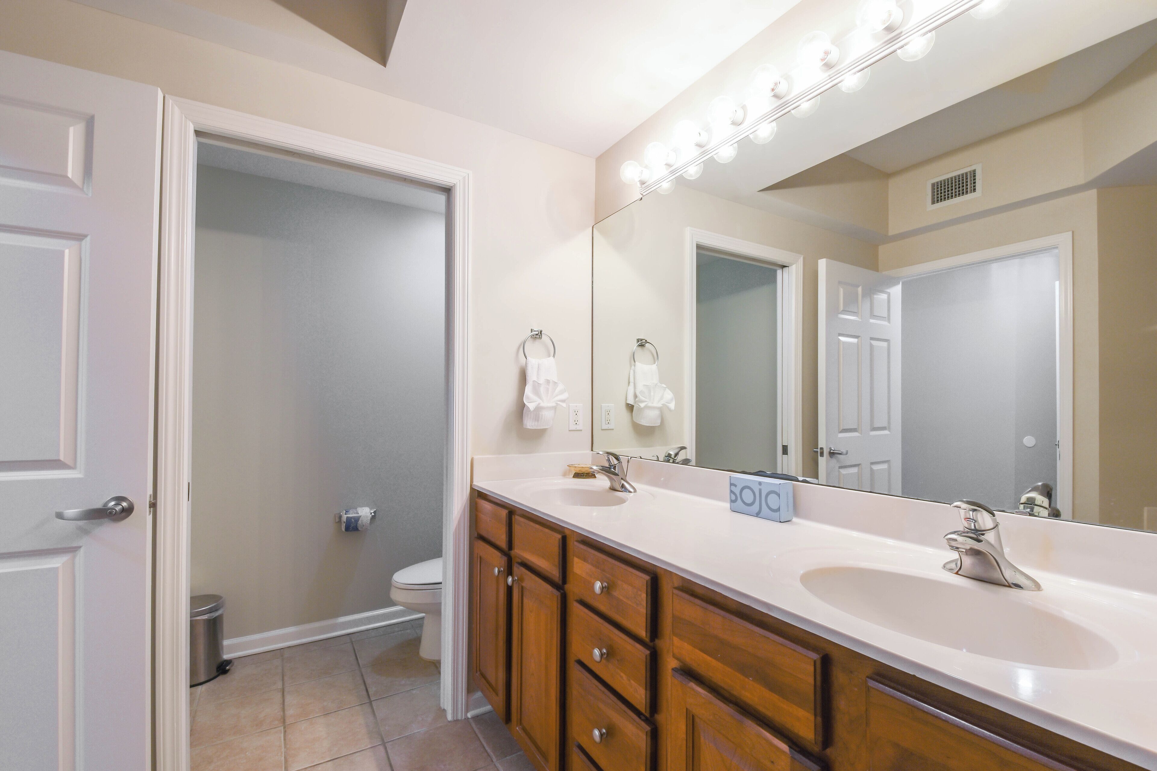 A bathroom with a vanity, sink, and mirror. There is a door leading to another room, a trash can, and a toilet.