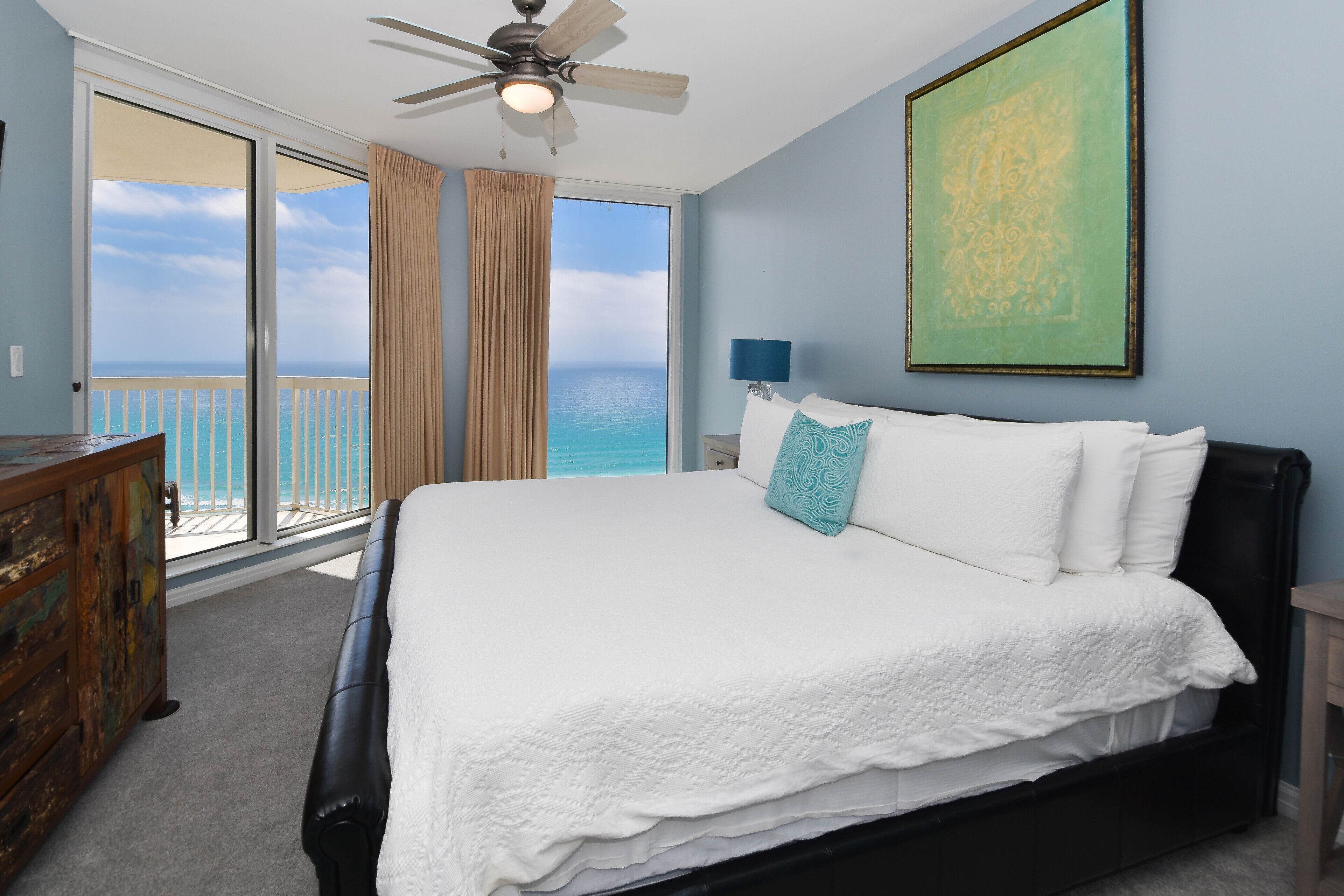 A bedroom with a large bed, a ceiling fan, a view of the ocean, and a painting on the wall.