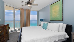 A bedroom with a large bed, a ceiling fan, a view of the ocean, and a painting on the wall.