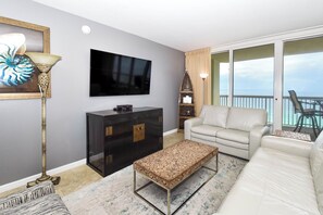 A living room with a flat-screen TV, a black entertainment unit, a beige sofa, a patterned ottoman, a floor lamp, and a view of the ocean through large sliding doors.