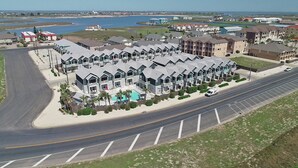 A coastal residential area with a row of modern houses featuring balconies, a swimming pool, and a road with a parking lot. - Pet-Friendly Beach Escape | Heated Pool & Private Patio (Corpus Christi)
