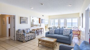 A spacious living room with a blue sofa, patterned armchairs, a coffee table, and a view of the ocean through large windows. - Gulf Front Condo, Elevator, Near Shops and Restaurants-Dune Allen Beach (Santa Rosa Beach)